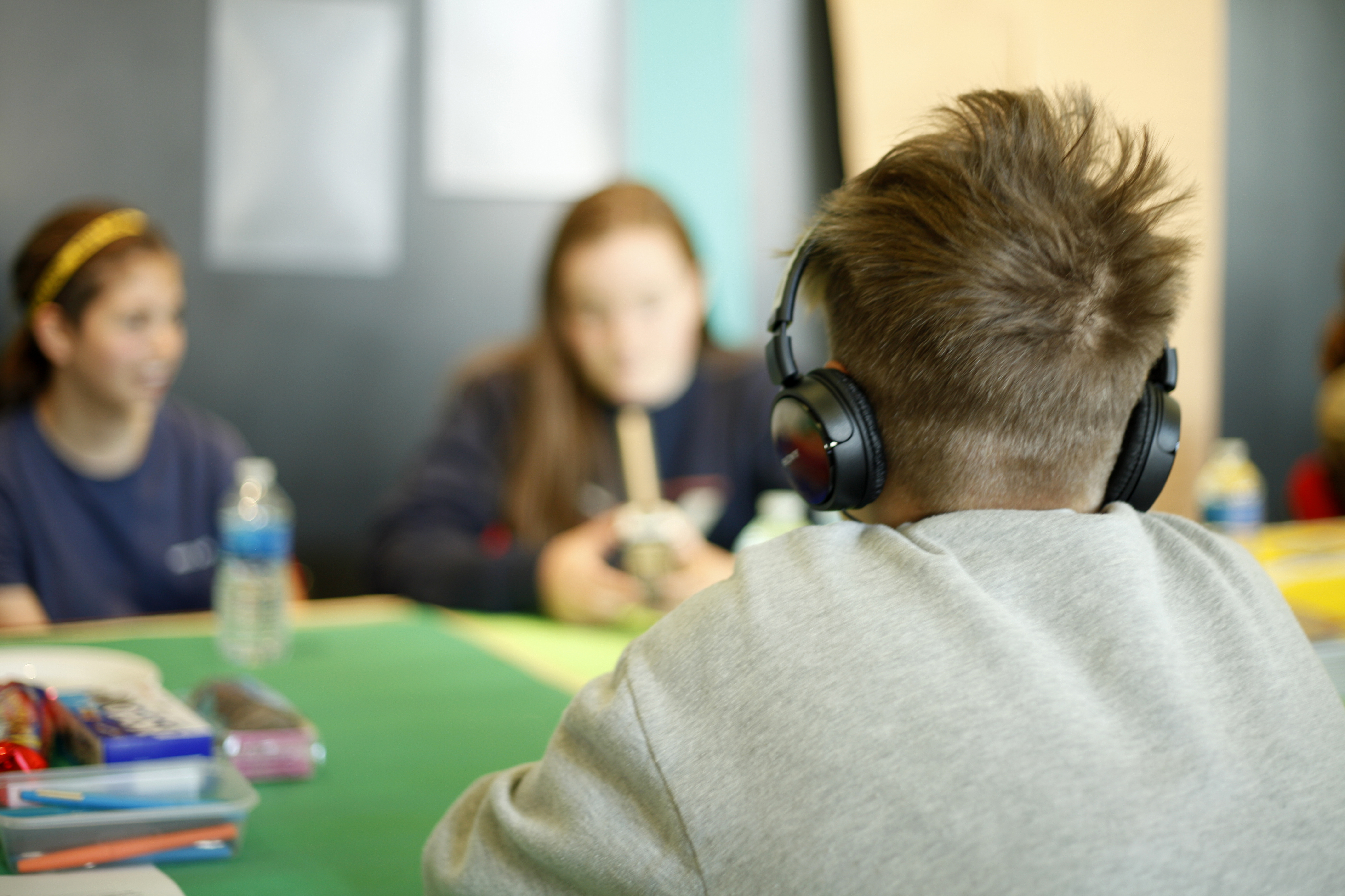Two young women sit opposite each other during the creation of a podcast, the one nearest to the camera has their back to us and is wearing headphones