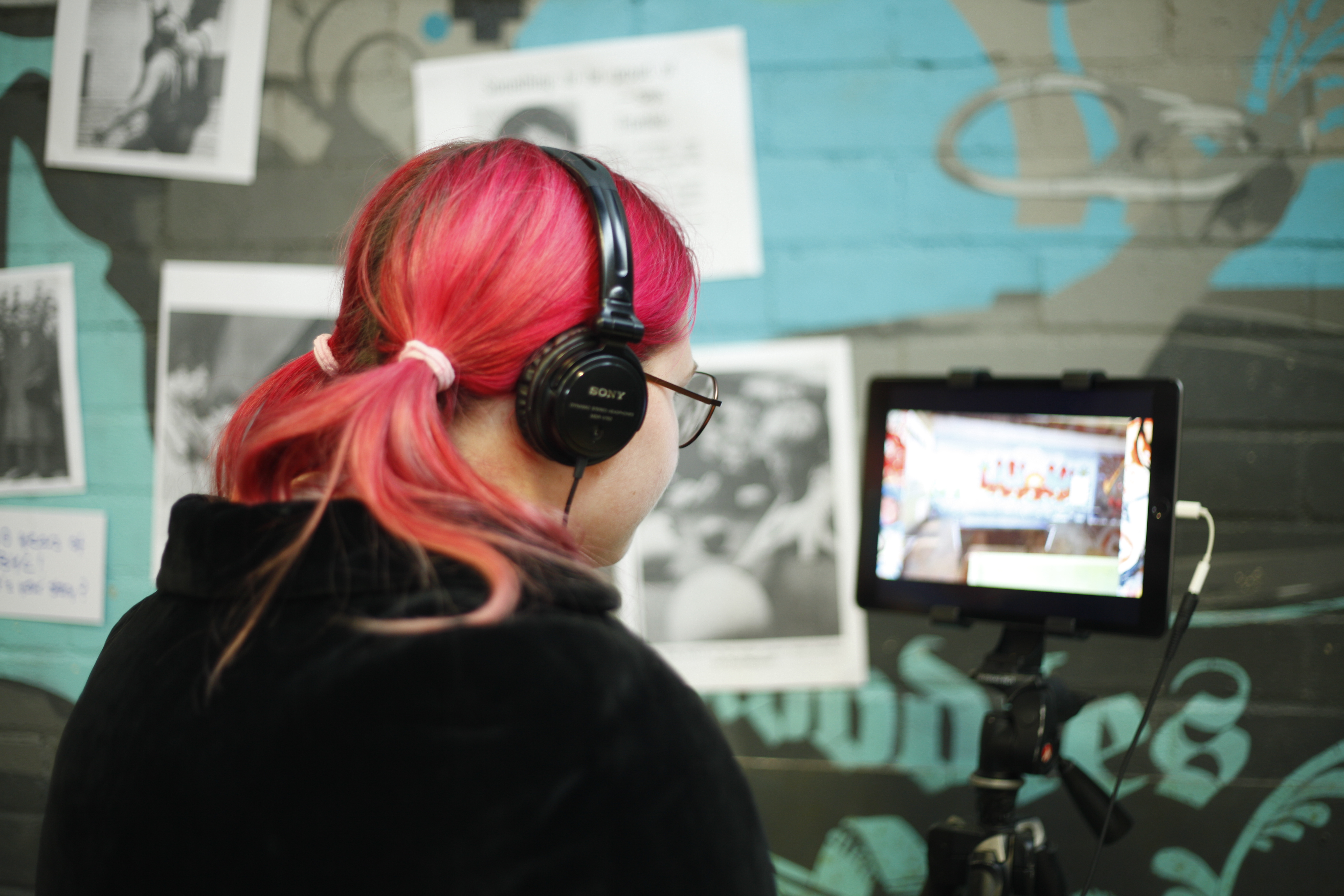 A young woman with pink hair is wearing headphones whilst watching a video on an iPad at the Youth Festival Launch at Brighton Youth Centre